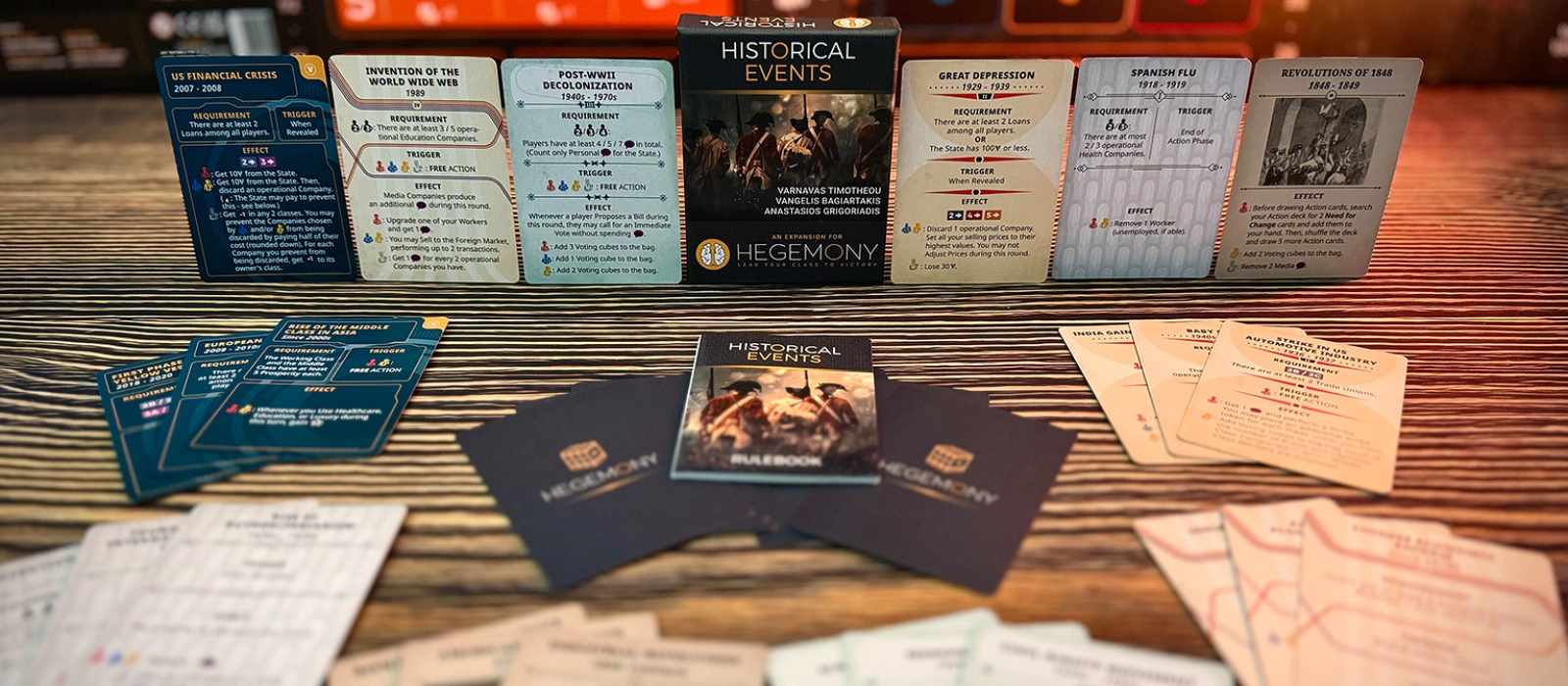 Hegemonic: Historical Events Expansion components displayed on a table. The game box stands upright in the center, surrounded by fanned-out card sets and the rulebook.