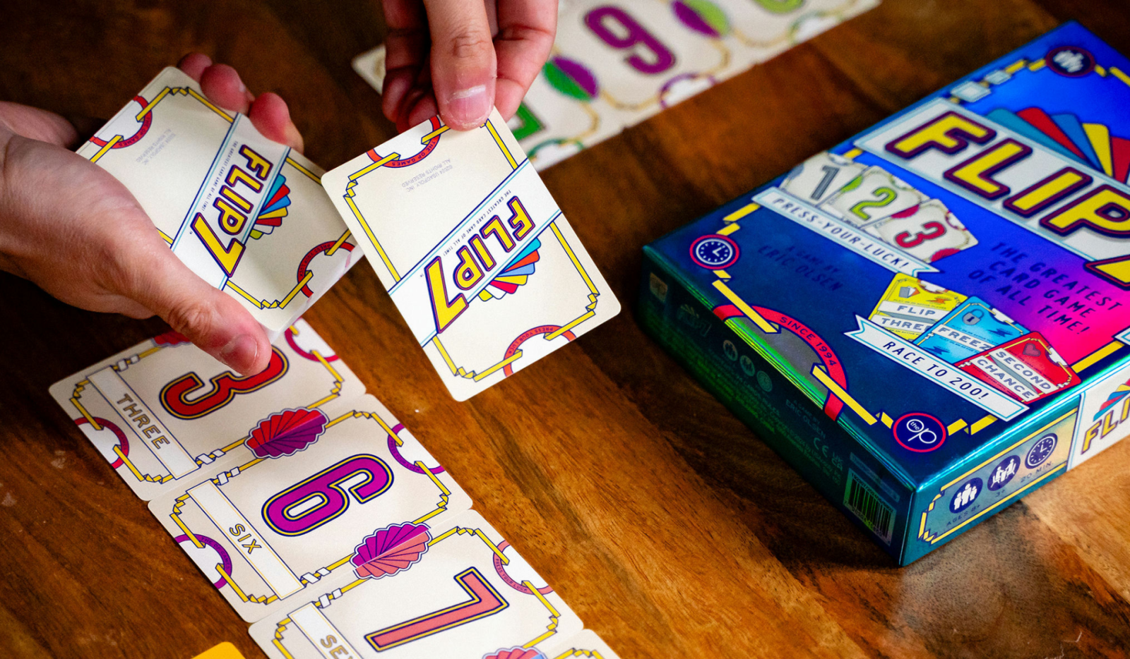 A lifestyle photo of the card game Flip 7 from the Op where someone is holding 2 cards in his hand. There are cards on the table and the box of Flip 7 is also on the table.