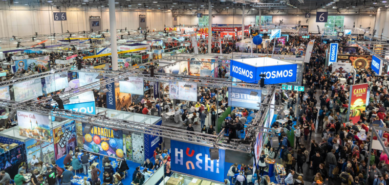 A high-angle view of SPIEL ESSEN, the world's largest board game convention, showcasing a crowded exhibition hall filled with attendees exploring booths, playing games, and engaging with publishers. Various exhibitor stands, including Kosmos, Catan, and other board game brands, are visible, with banners, demo tables, and vibrant displays creating a lively atmosphere.