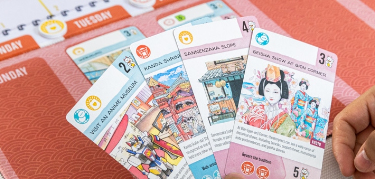 A close up from a hand with the cards of the board game Let's Go! To Japan with a part of the board game in the background. From the publisher The Op (USAopoly).